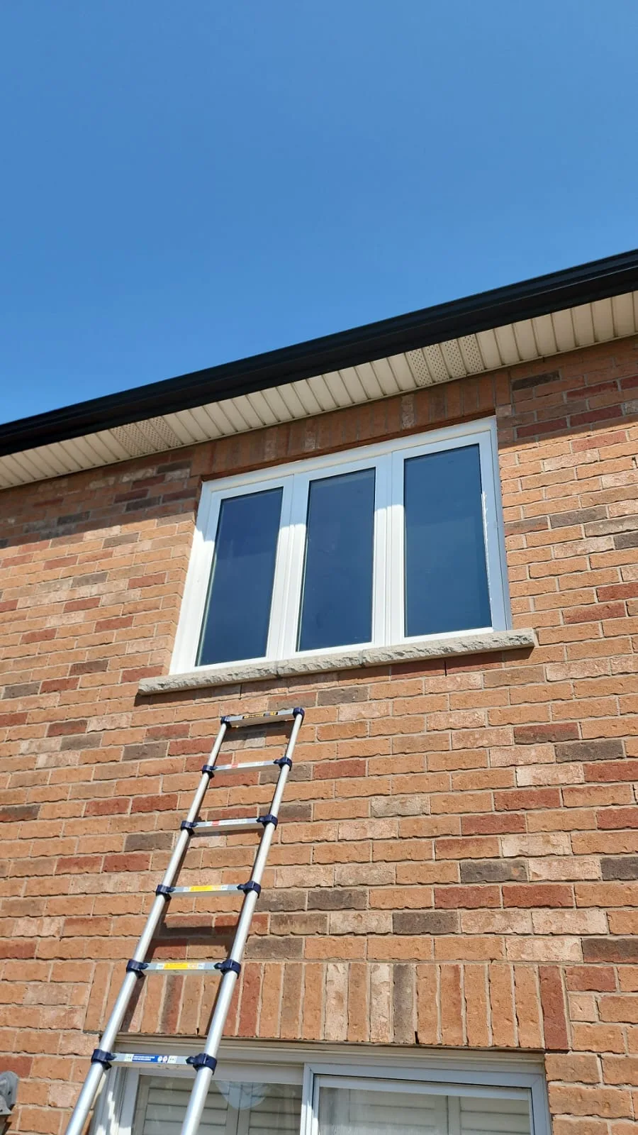 Three-panel white window on the upper floor of a brick house, with a ladder still in place below